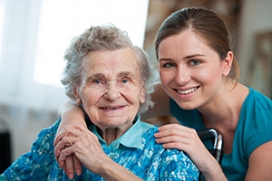 Senior woman sitting down and smiling wearing a blue and green sweater with a young woman crouched down behind her with her hands on the senior's shoulders