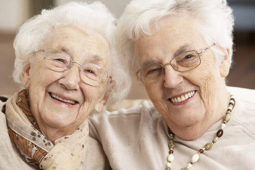 Two senior woman smiling with glasses on, one wearing a scarf and one wearing a necklace