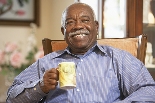 Senior man with a mustache sitting in a chair holding a coffee cup and smiling